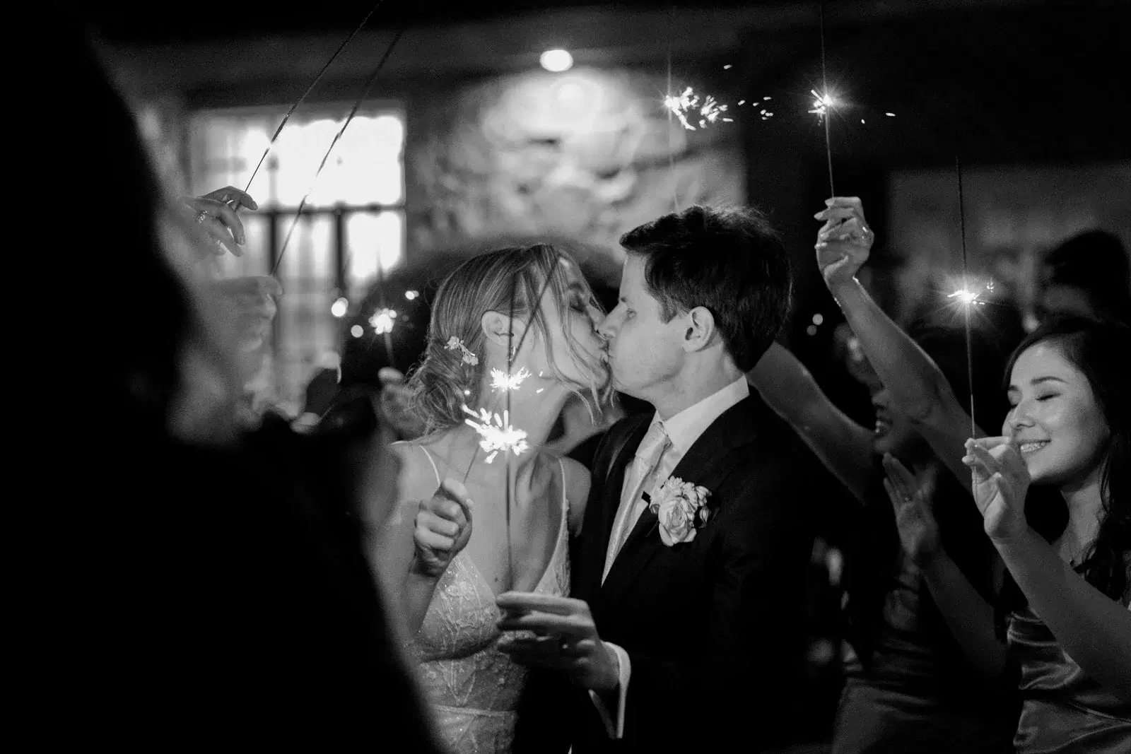 Newlyweds kissing while guests celebrate with sparklers